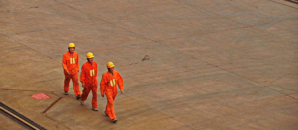 Three workers in hi-vis overalls and hard hats walking across a wide, empty concrete apron